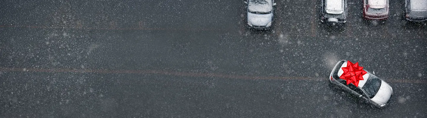 Aerial view of snowy parking lot with several cars; one silver car dusted in snow sports a large red gift bow on its roof.