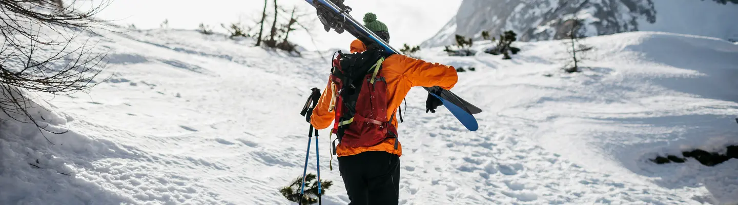 Back view of a skier in an orange jacket and green beanie carrying skis and poles uphill through snowy mountain terrain.