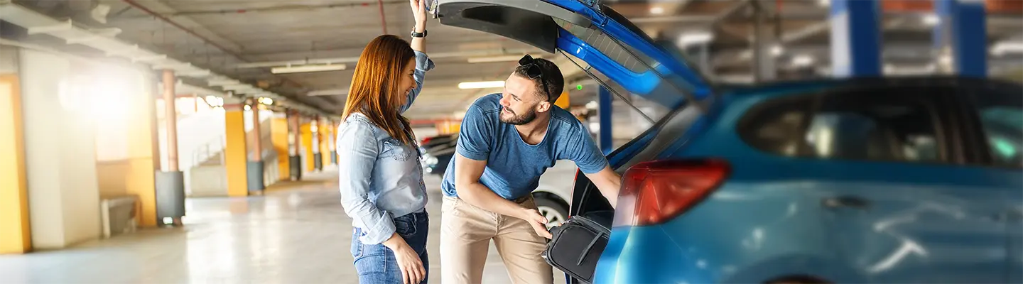 A man and woman talk while loading a suitcase into the trunk of a blue car in a parking garage.