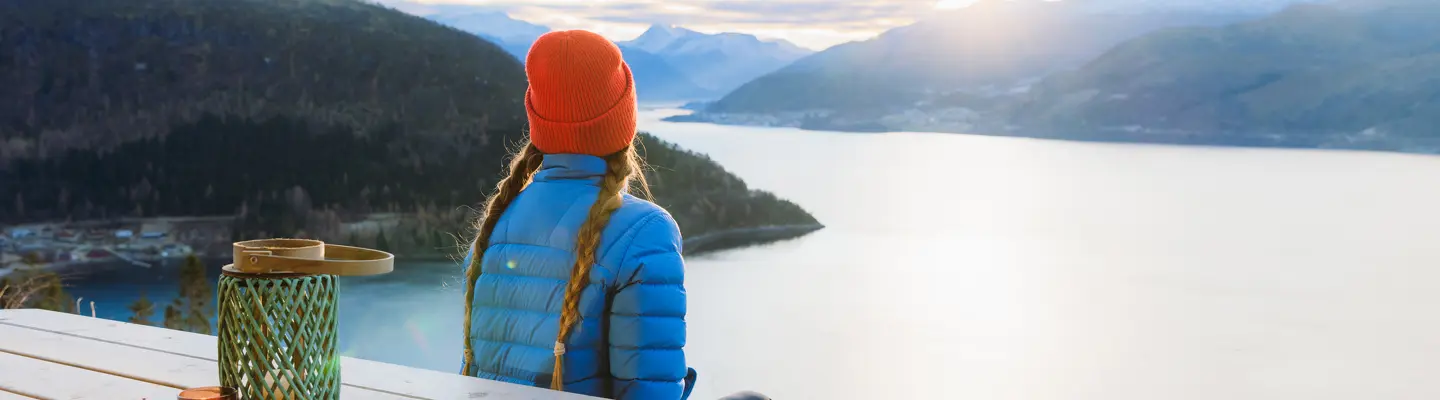 Person in red beanie and blue puffer jacket with braided hair sits on a deck overlooking sunlit fjord and mountains.