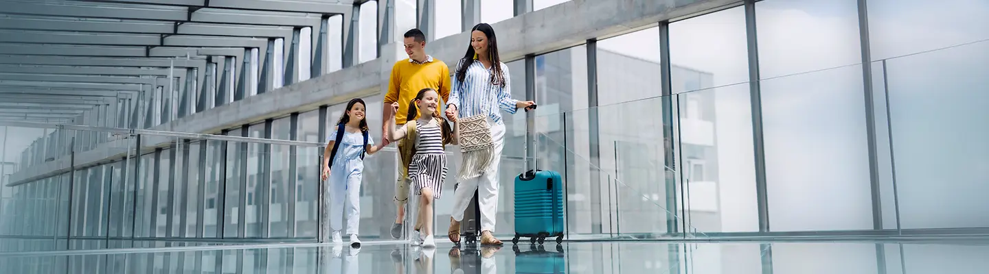 A family of four, with two young girls, walks through an airport terminal, pulling luggage. The terminal features glass walls and a reflective floor.