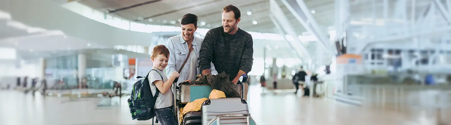 A family of three smiling and pushing a luggage cart in a bright, spacious airport terminal.
