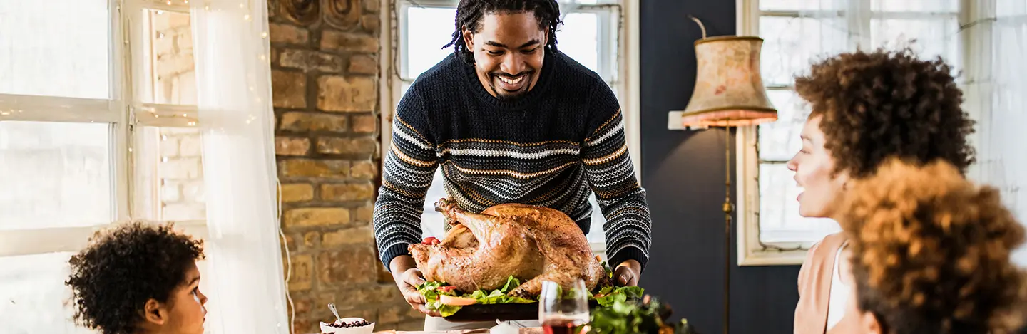 A person places a roasted turkey on a table surrounded by family members, with warm lighting and a cozy atmosphere.