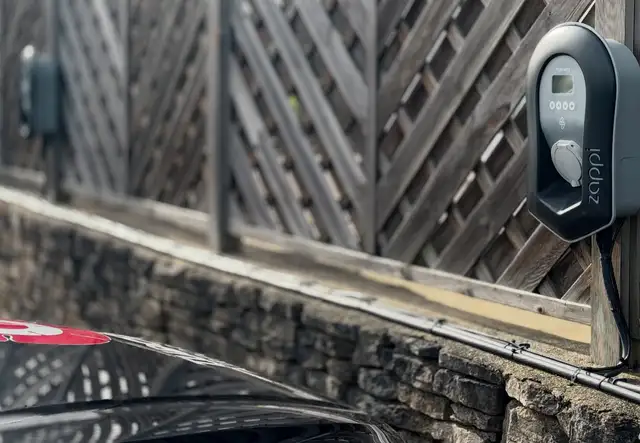 Electric vehicle charger mounted on a wooden fence, with a black car partially visible in the foreground.