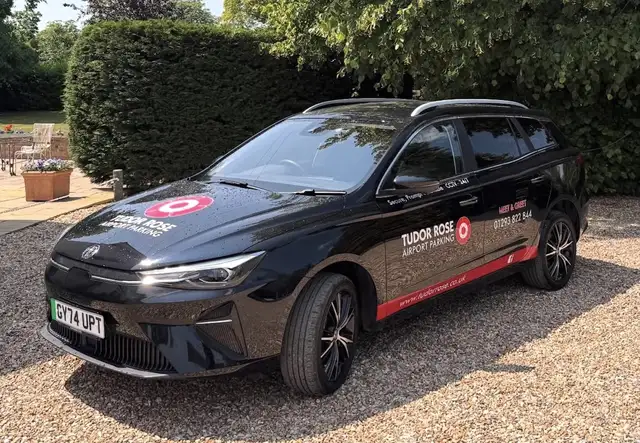 Black Tudor Rose Airport Parking car on gravel driveway, with business signage and contact details, surrounded by greenery.