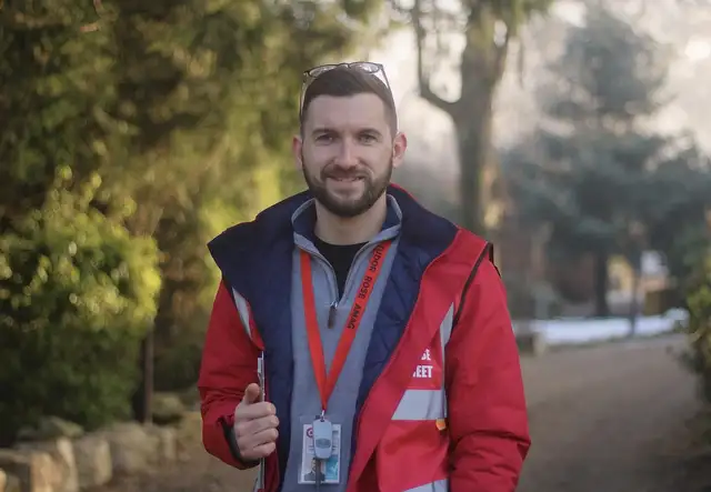 Man in a red jacket with a badge and lanyard stands on a path surrounded by greenery, holding a pen and paper, smiling at the camera.