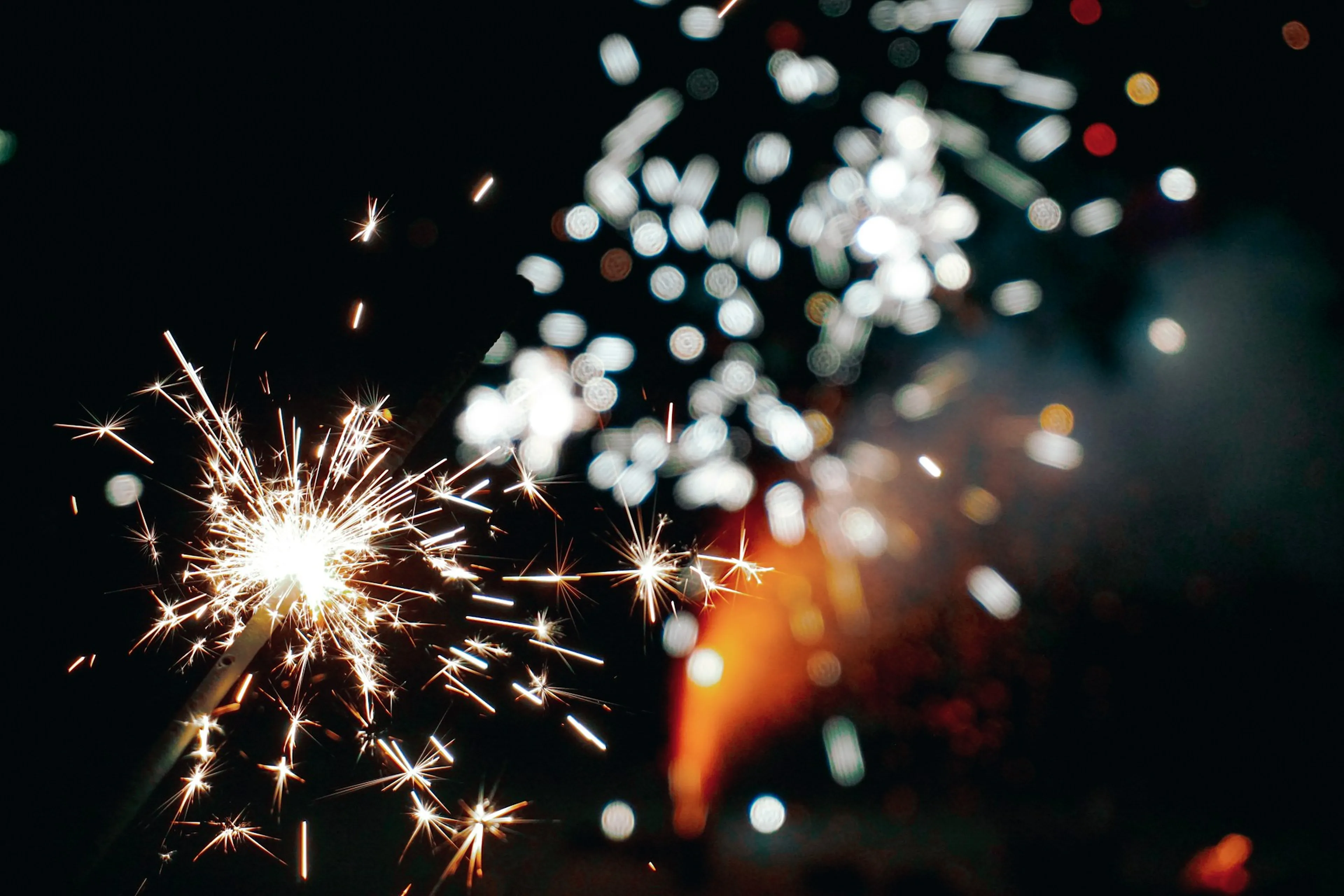 Close-up of bright sparklers emitting sparks against a dark background, with shimmering lights in the distance.