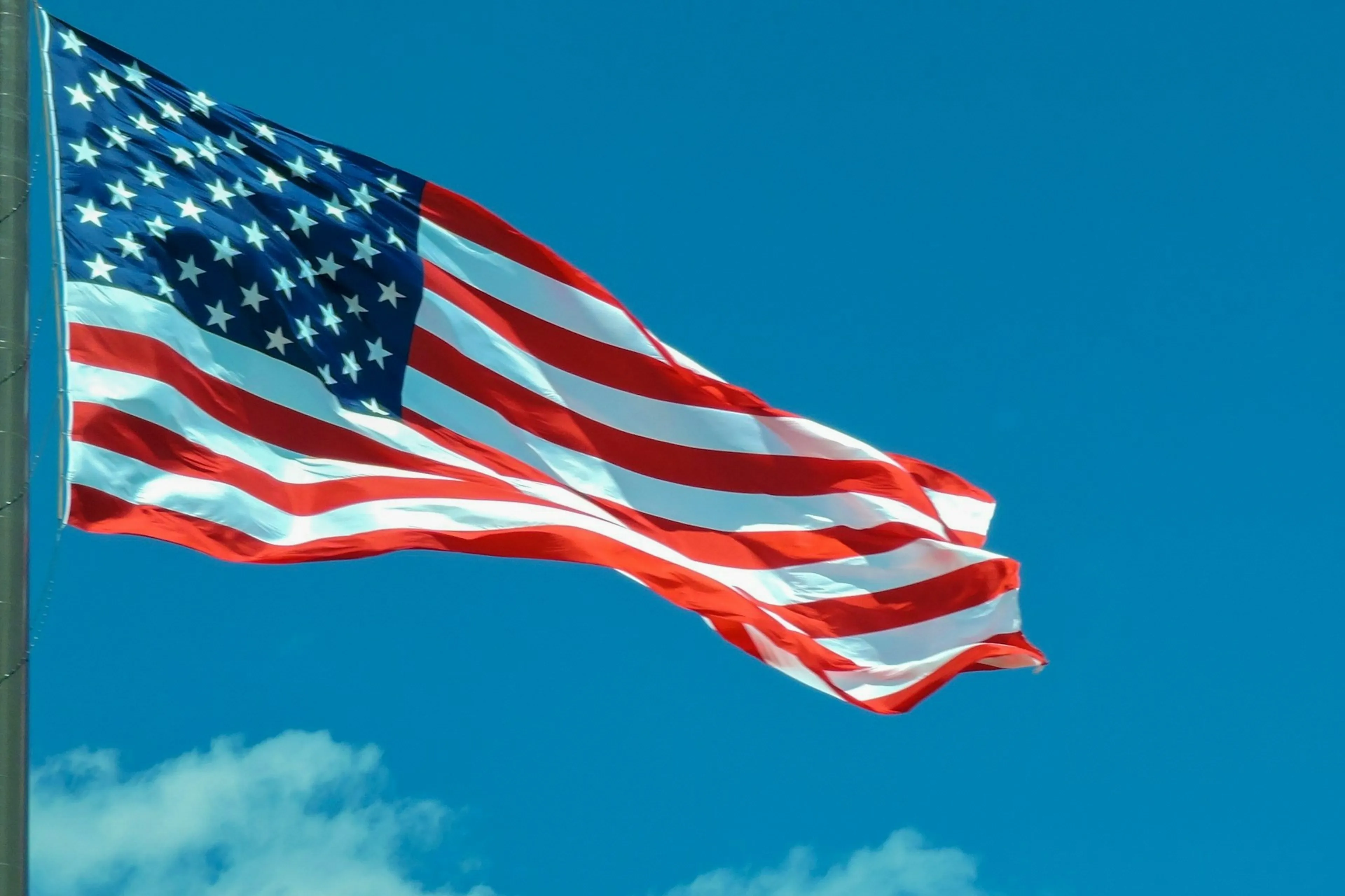 The American flag waves against a clear blue sky, displaying red and white stripes with a field of white stars on a blue background.