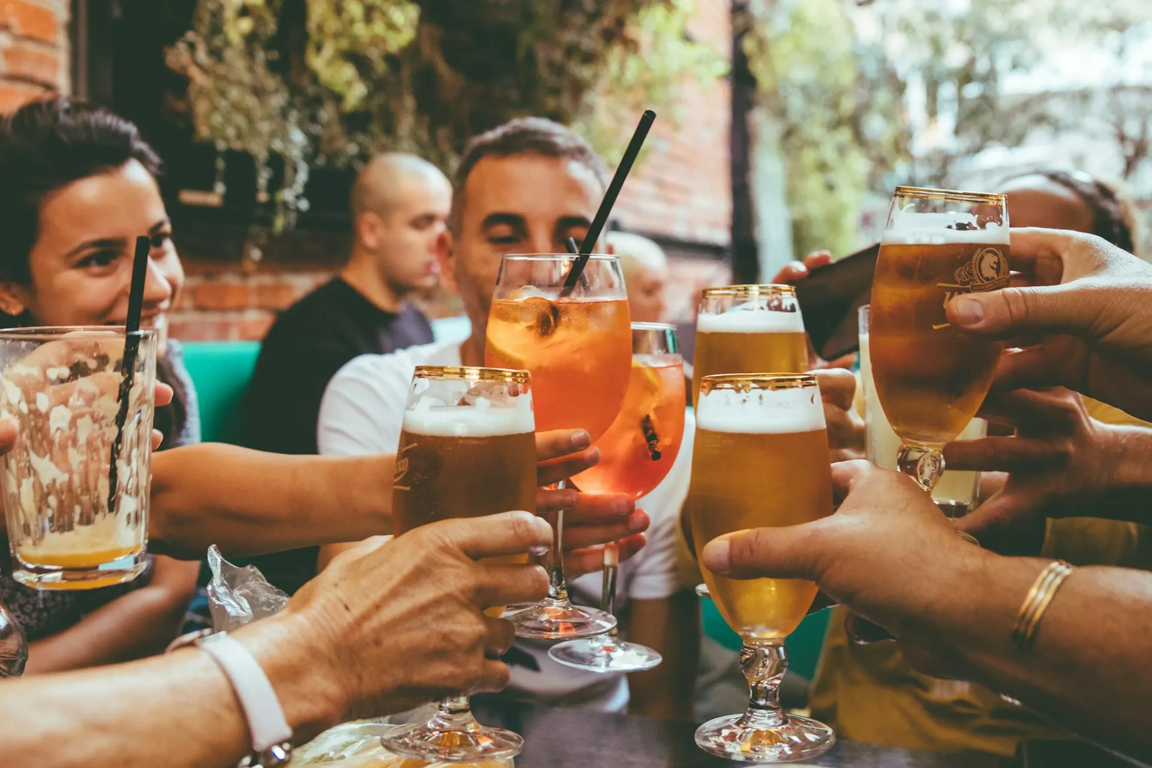 People toasting with various drinks, including beer and cocktails, in an outdoor setting with brick walls and greenery.