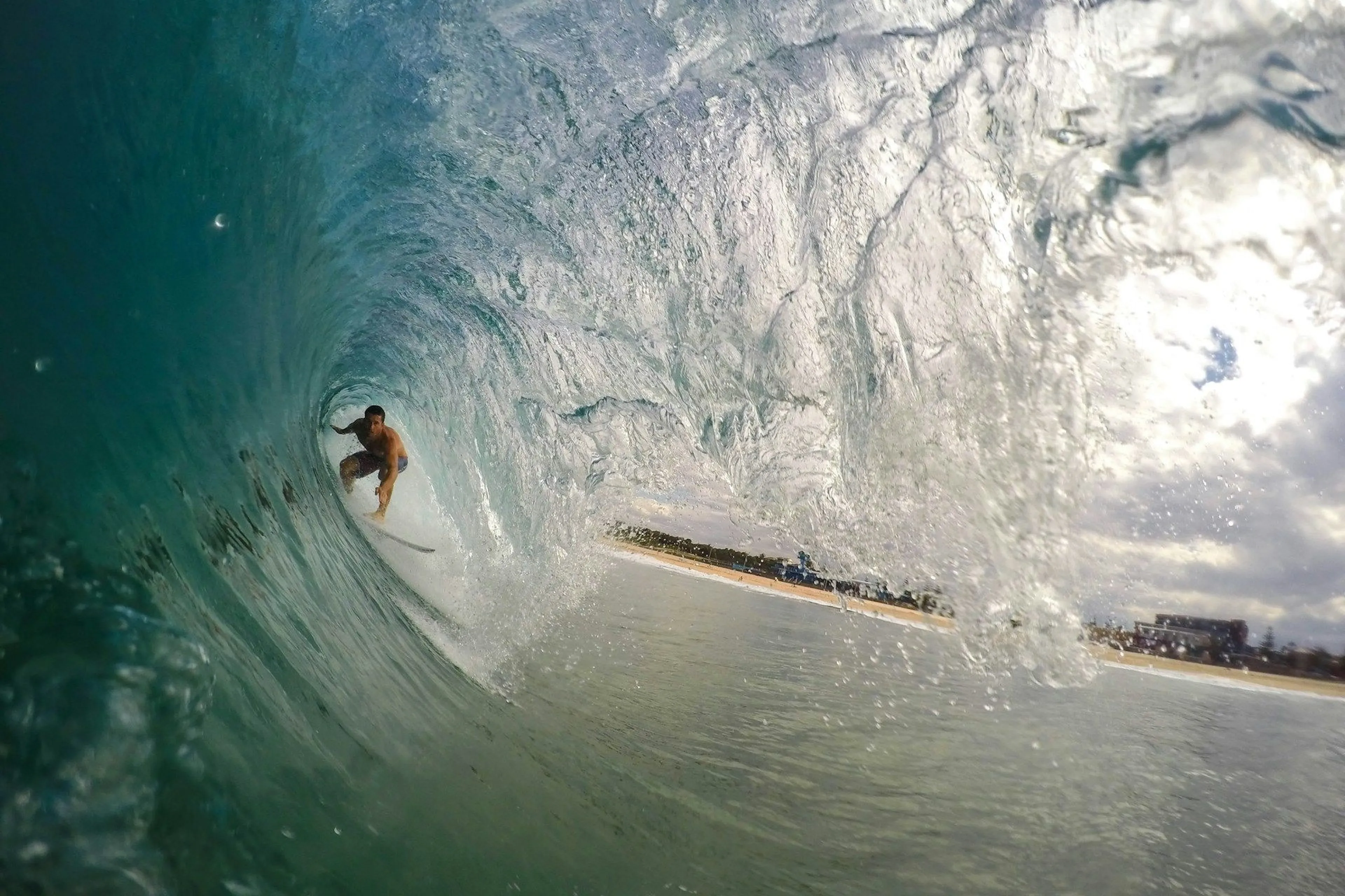 Surfer riding inside a large, breaking ocean wave near the shoreline under a cloudy sky.