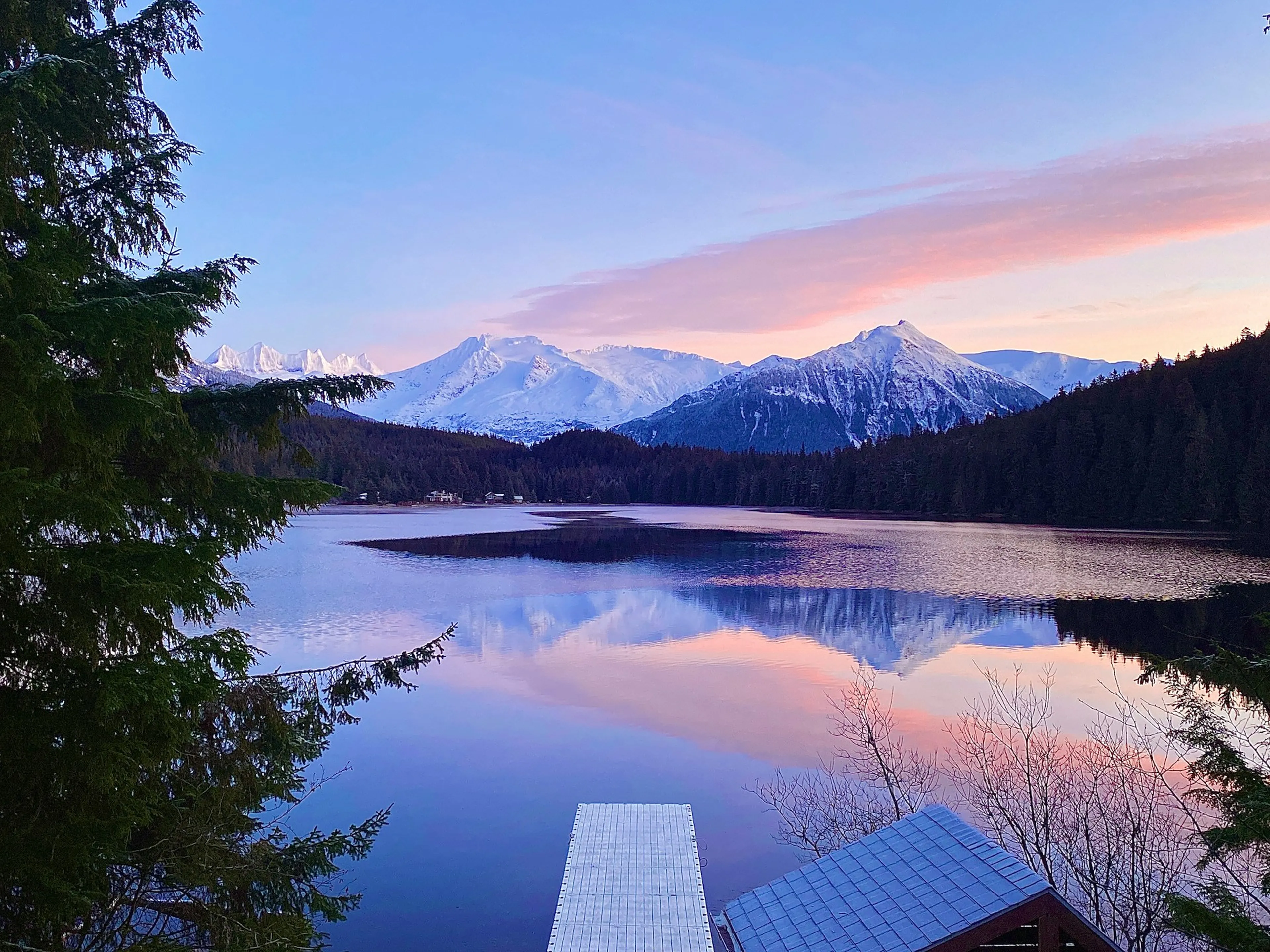Snow-capped mountains and pink sunset sky reflected on a serene lake, surrounded by evergreen trees and a small dock in the foreground.