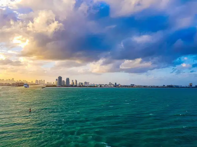 City skyline at sunset with tall buildings, vibrant blue-green ocean, and dramatic clouds in the sky.