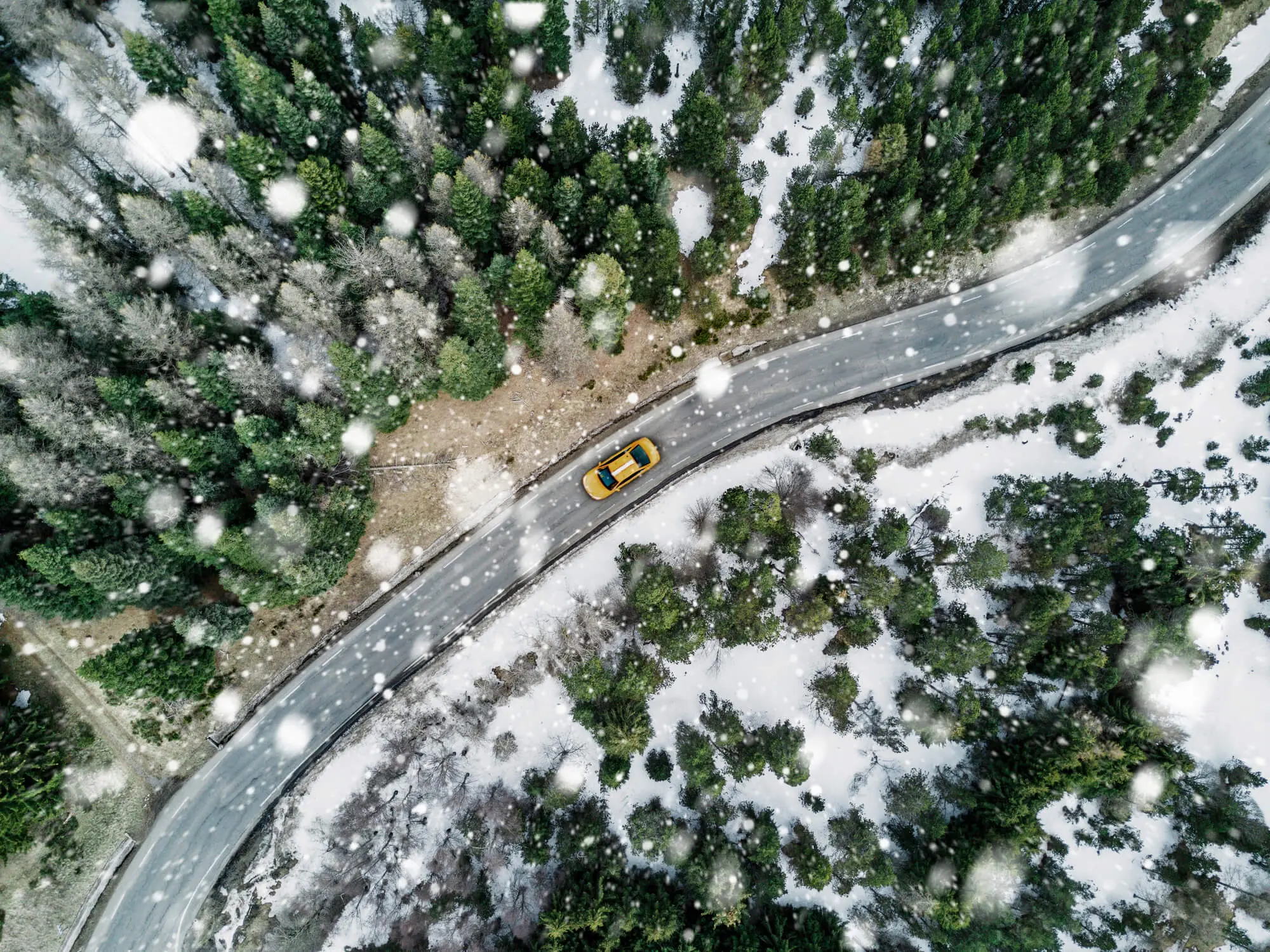 Aerial view of a yellow car driving on a snowy, winding road through a forest, with snowflakes falling around.