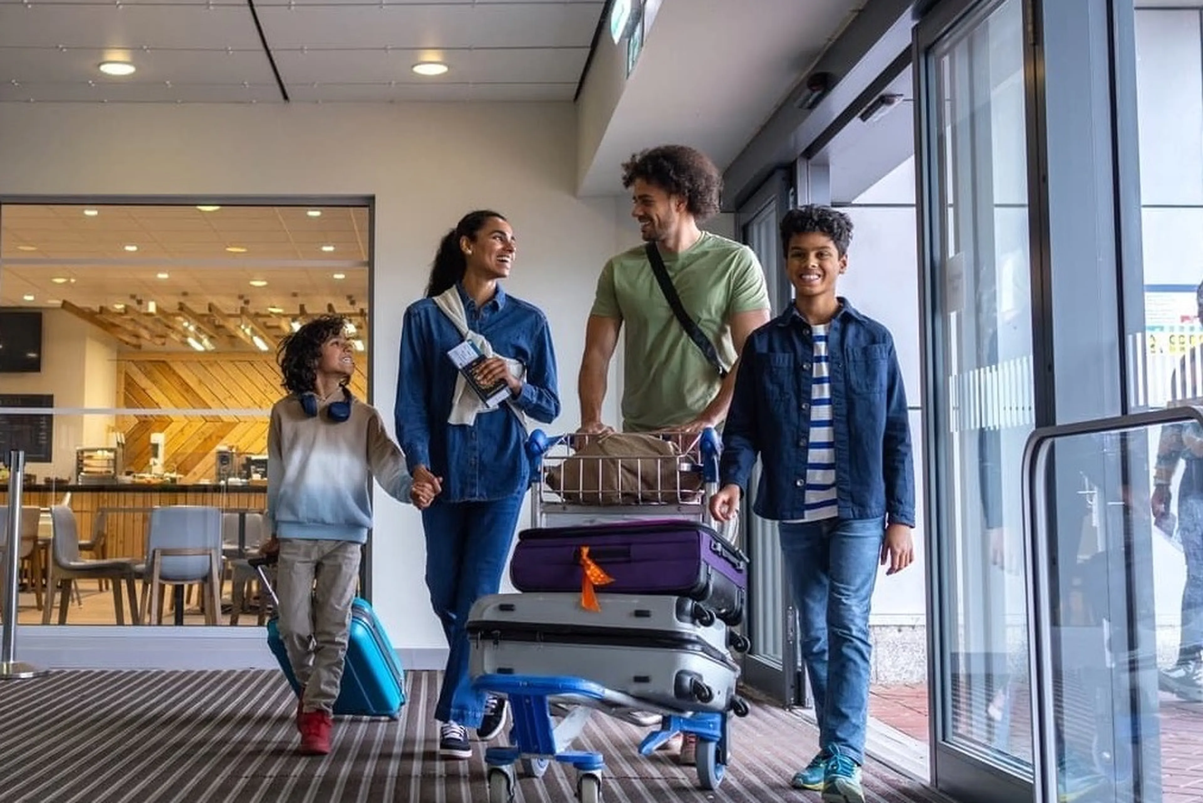 A family of four walking through an airport entrance, smiling and carrying luggage on a trolley.