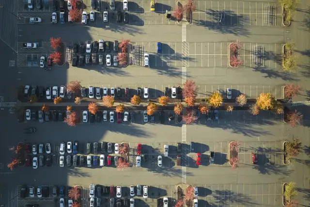 Aerial view of a parking lot with rows of parked cars and autumn trees casting long shadows in the sunlight.