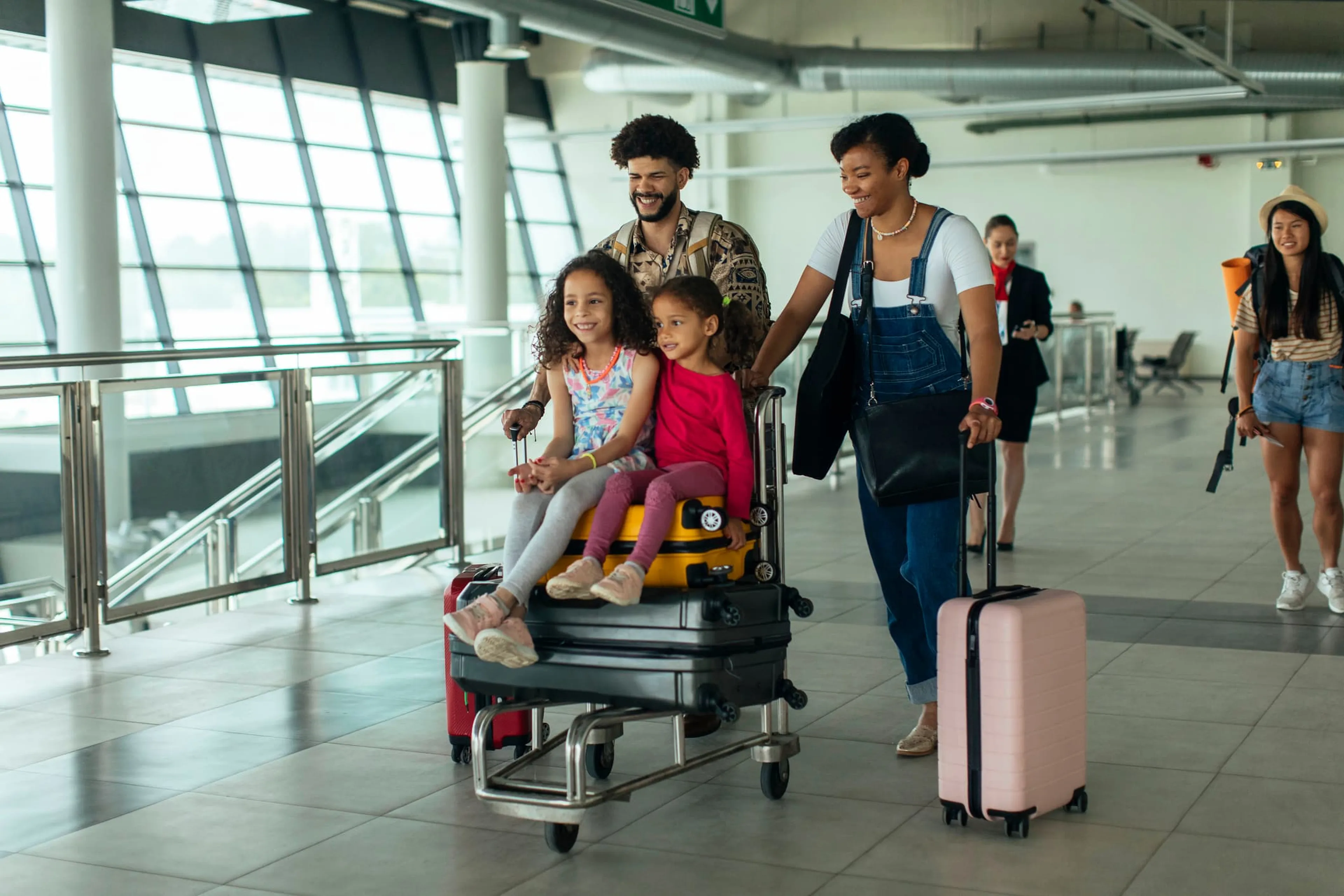A smiling family with two children on luggage carts at an airport terminal, surrounded by travelers with suitcases.