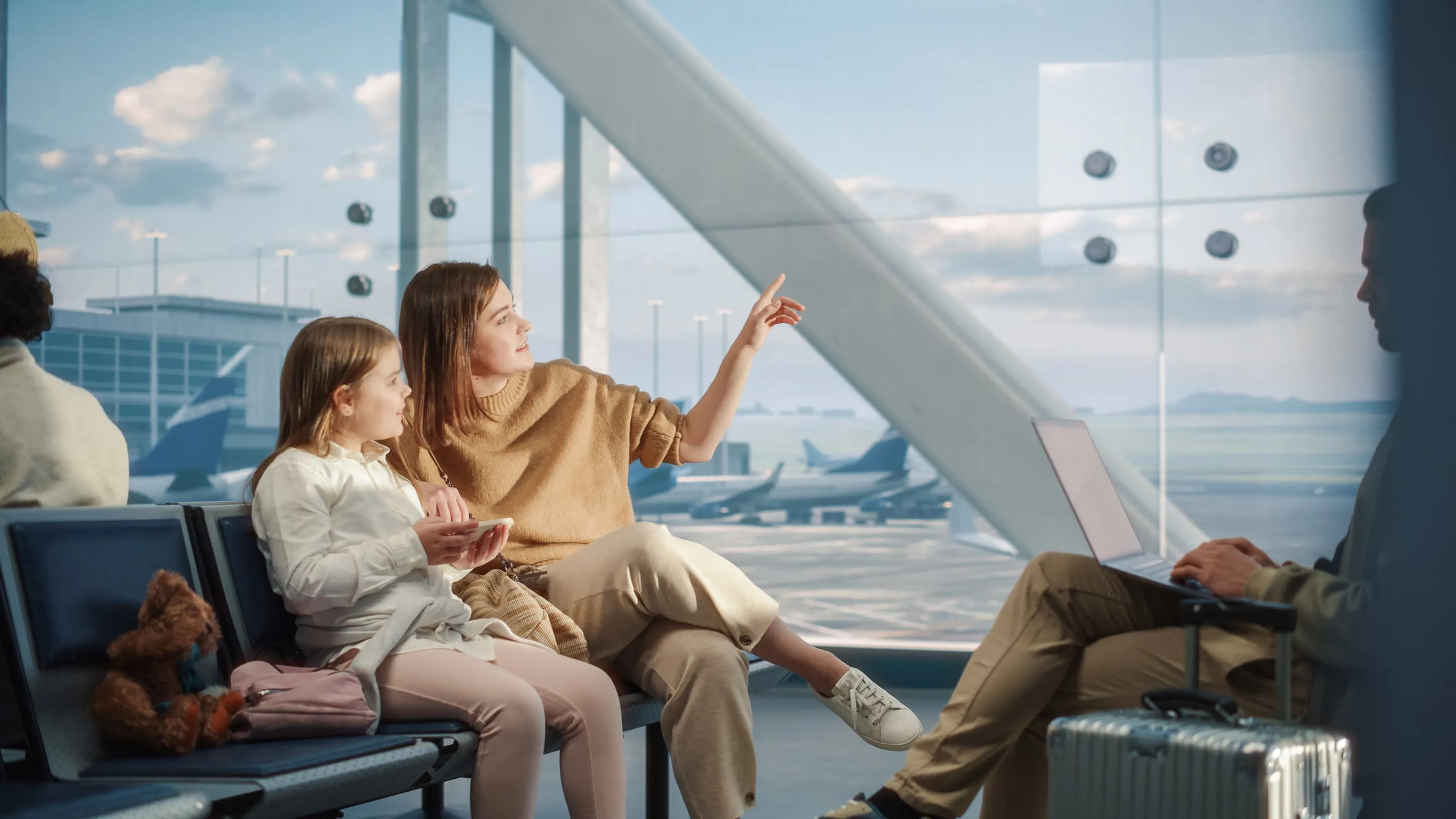 A woman and a child sit on airport seats while the woman points outside. A teddy bear and bag are beside them. A man works on a laptop in the background.