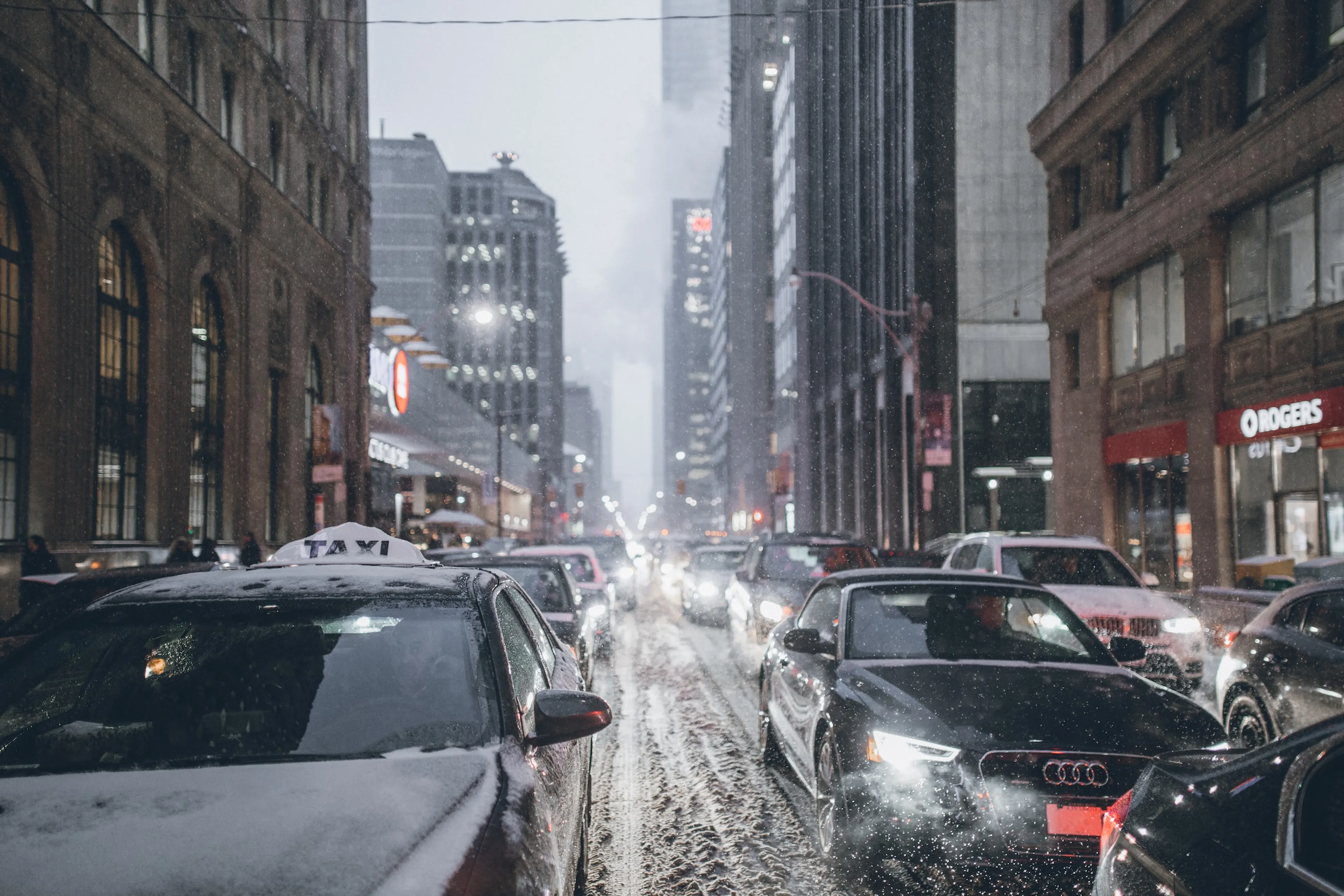 Snowy city street with heavy traffic, surrounded by tall buildings. Cars are lined up, headlights glowing in the dim, wintry atmosphere.
