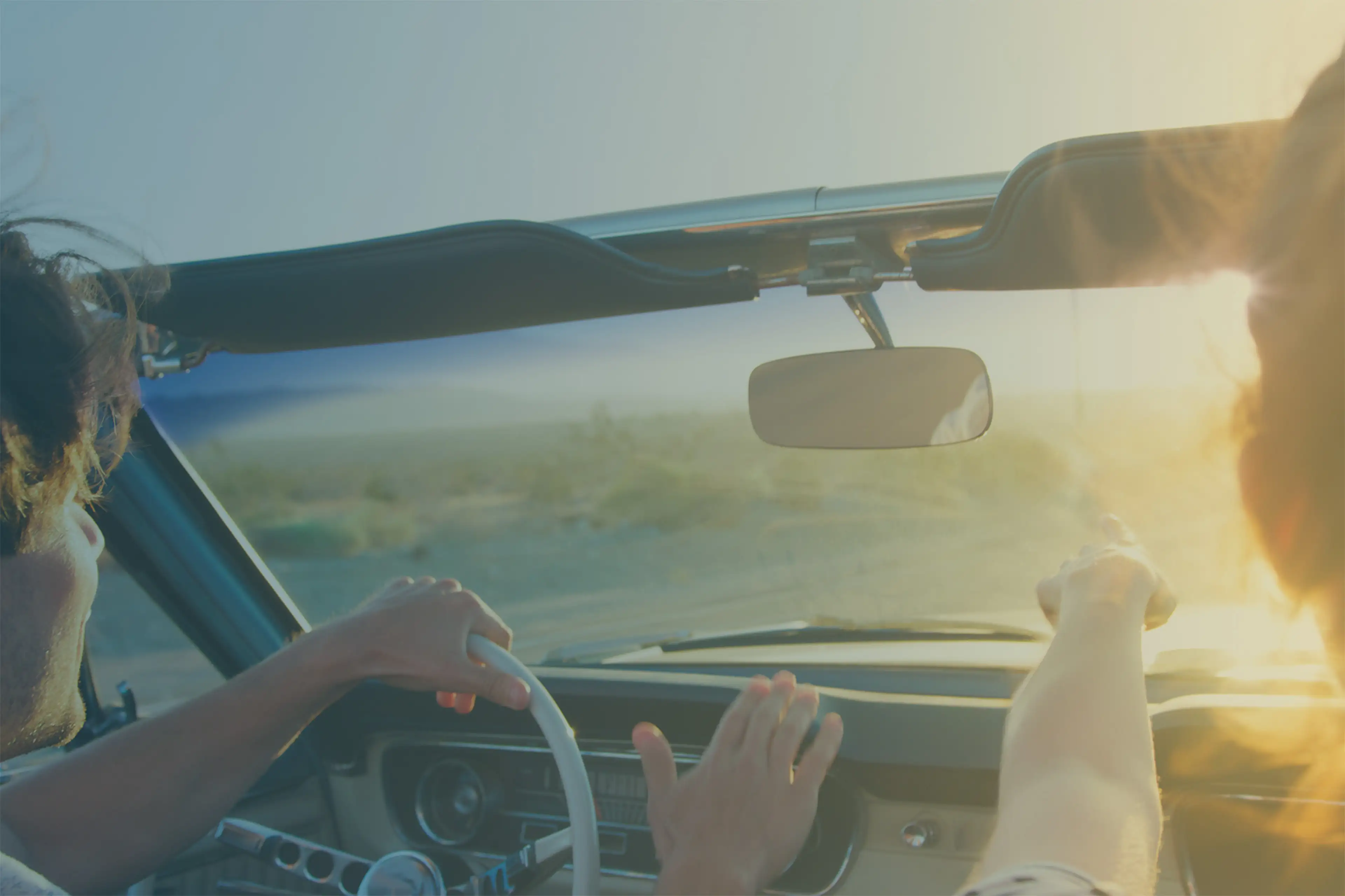 Two people in a vintage convertible drive through a sunlit desert landscape, one pointing ahead.