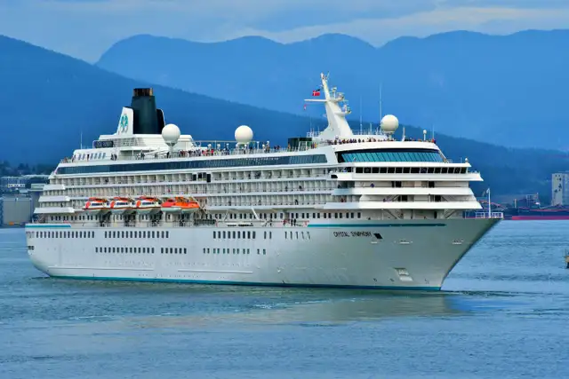 A large white cruise ship sails on calm waters with a mountainous backdrop under a cloudy sky.