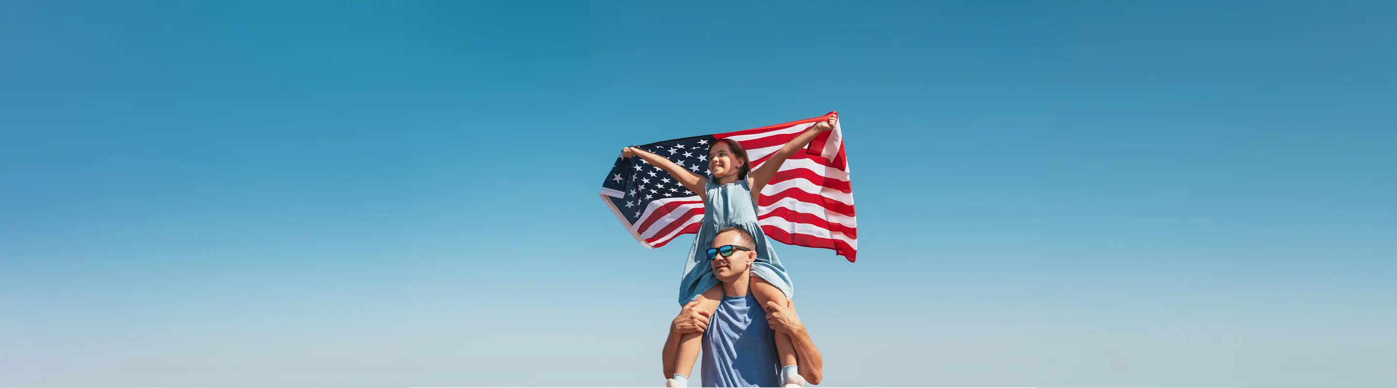 Child on an adult's shoulders holding a U.S. flag, against a clear blue sky, symbolizing freedom and patriotism.