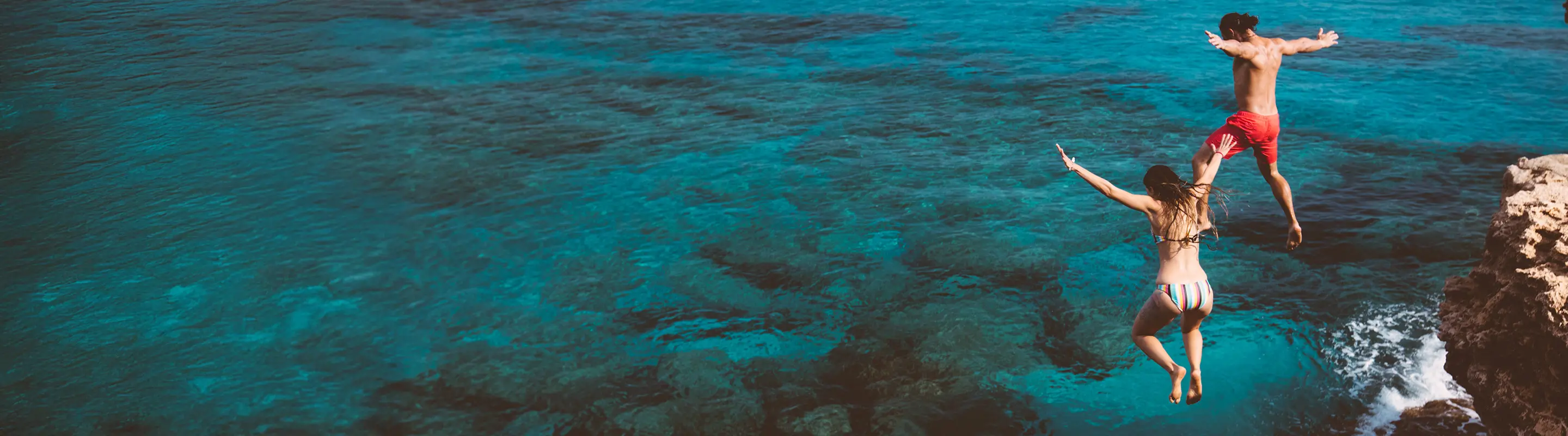 Two people in swimwear mid-air, jumping off a rocky cliff into clear turquoise water, with their arms outstretched.