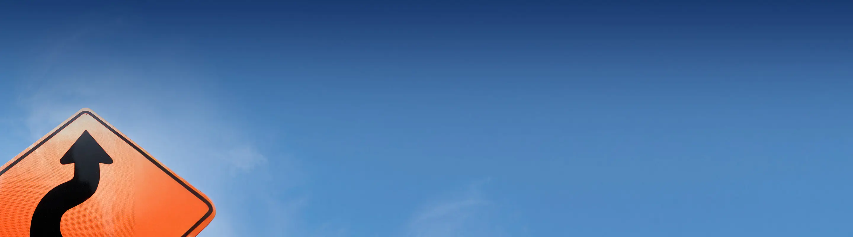 A yellow road sign with a curving arrow on the left side against a gradient blue sky background.