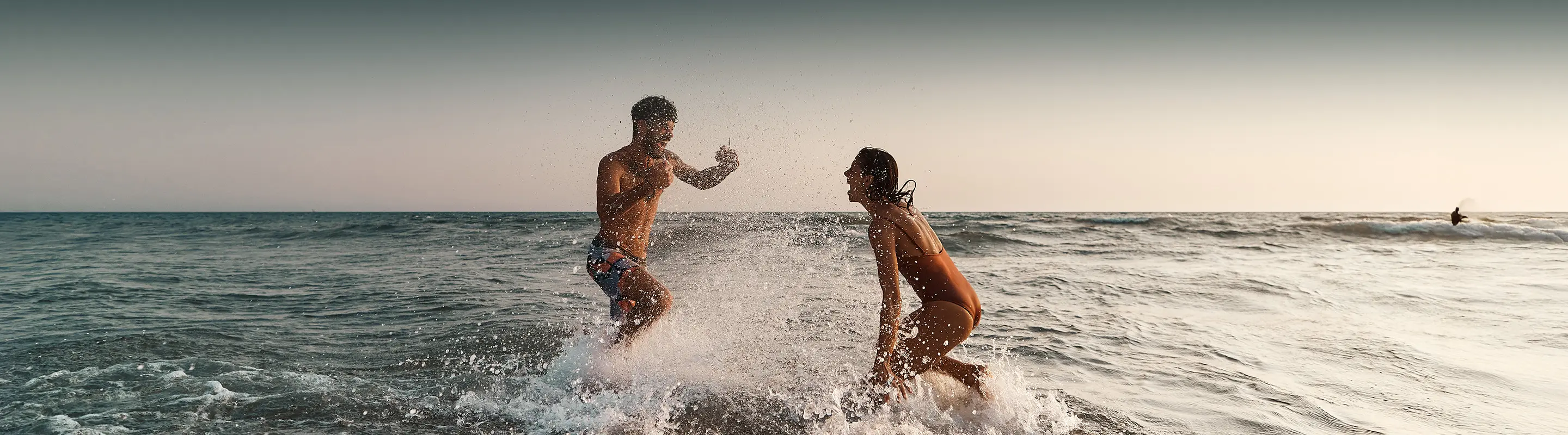 Two people splashing water at each other in the ocean at sunset, with waves in the background.