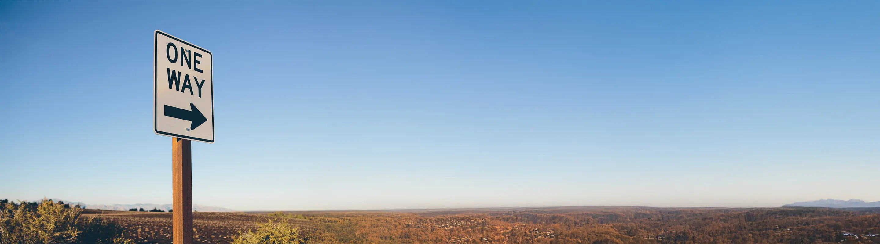 A "One Way" sign with an arrow pointing right stands against a clear blue sky and a vast, dry landscape.