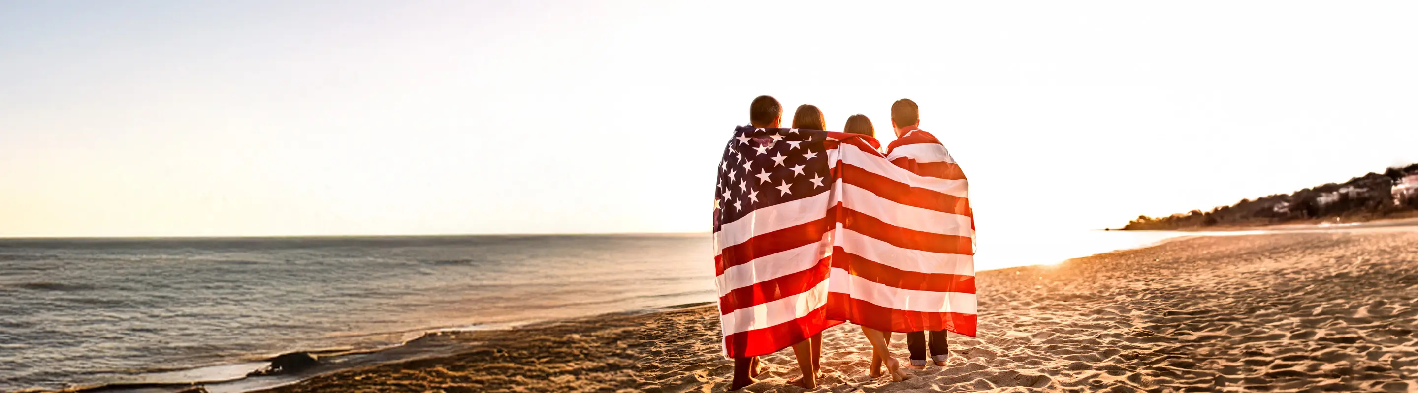 Four people wrapped in an American flag stand on a sandy beach, gazing at the ocean during sunset.