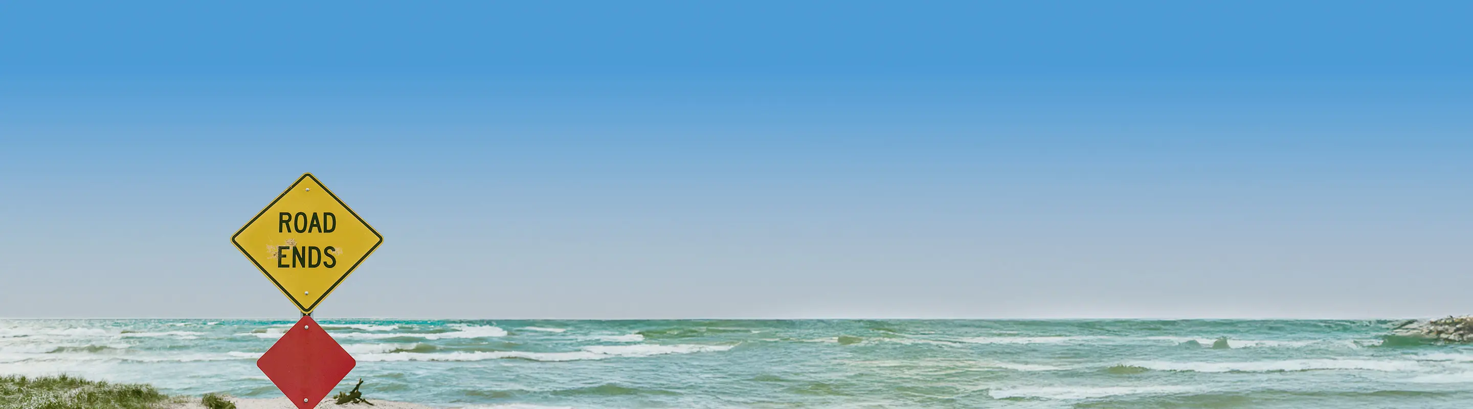 A "Road Ends" sign near a grassy shoreline with waves crashing under a clear blue sky.