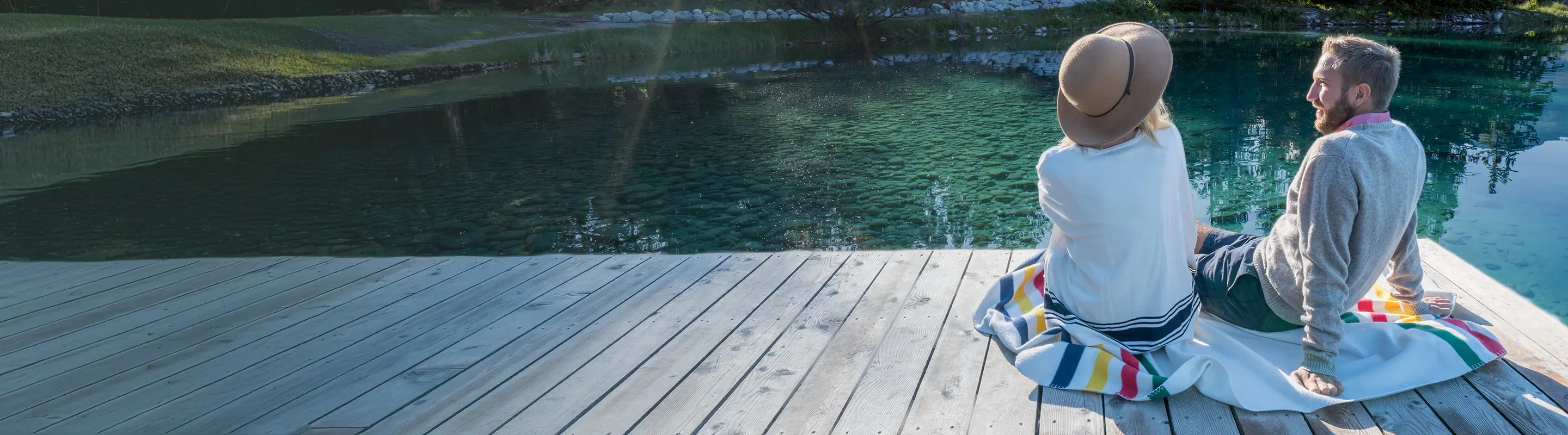 A couple sits on a wooden dock by a clear, tranquil lake, surrounded by greenery. They are relaxed, enjoying the serene view.