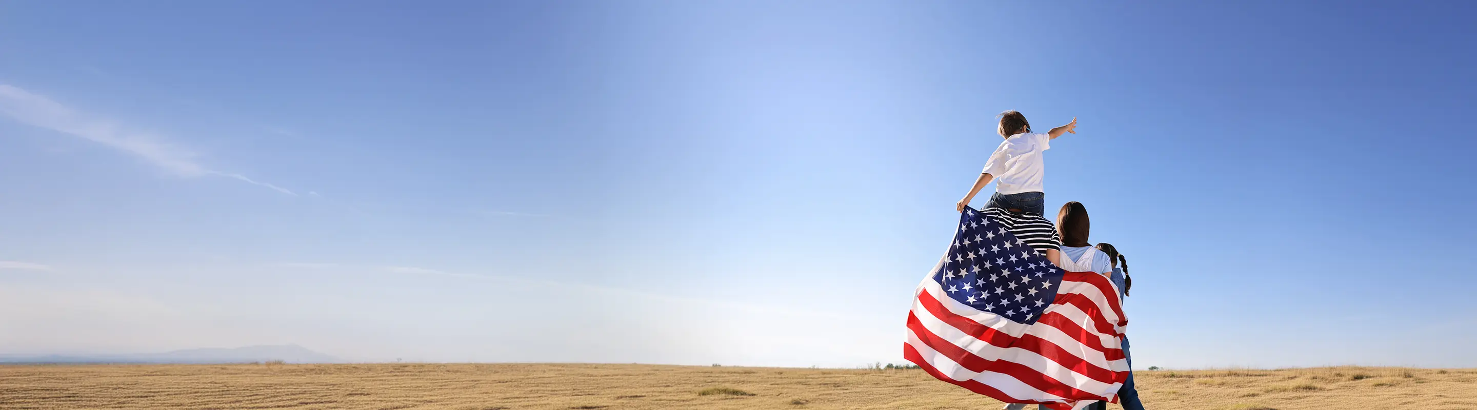 Child on adult's shoulders holding an American flag in a field under a clear blue sky, pointing towards the horizon.