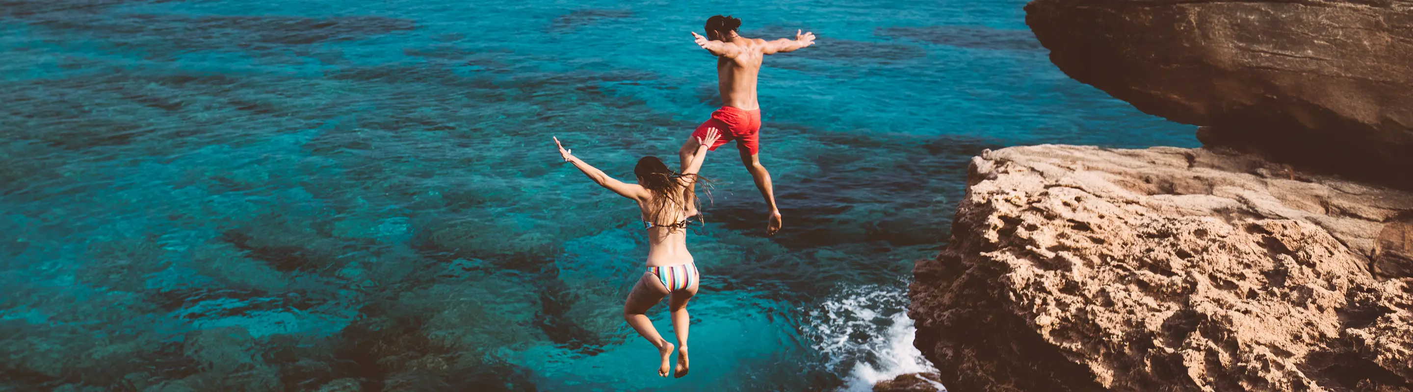 Two people in swimwear jump off a rocky cliff into clear blue ocean water, with waves crashing below.