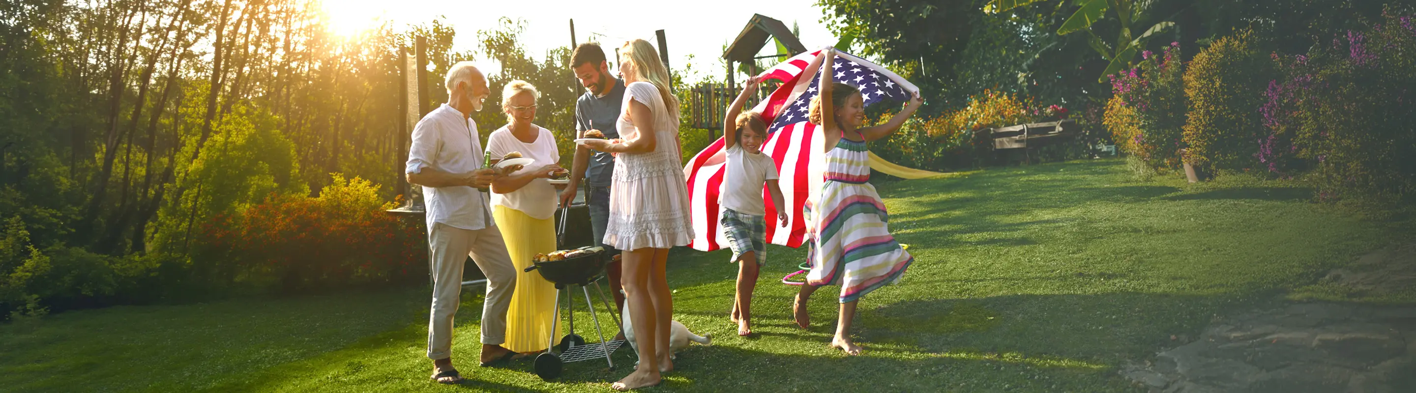 Family enjoying a barbecue in a sunlit garden, with children running carrying an American flag, surrounded by colorful flowers and greenery.