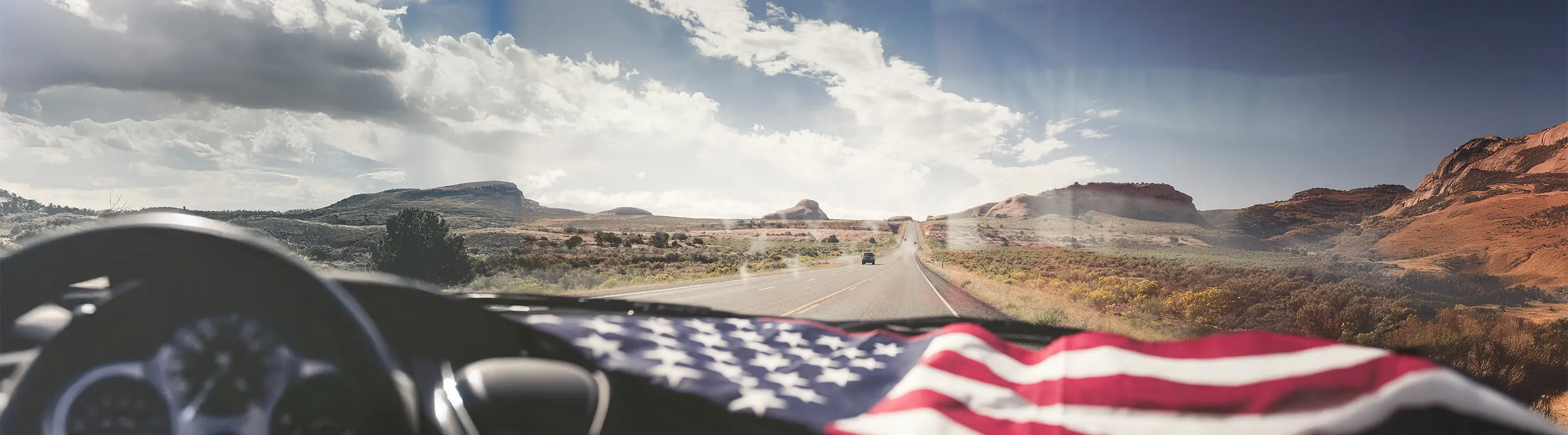 View from inside a car driving on a desert highway, with an American flag visible on the dashboard.