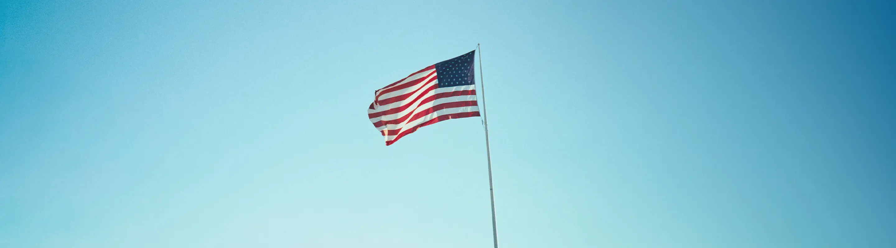 American flag waving against a clear blue sky, with sunlight illuminating the stars and stripes.