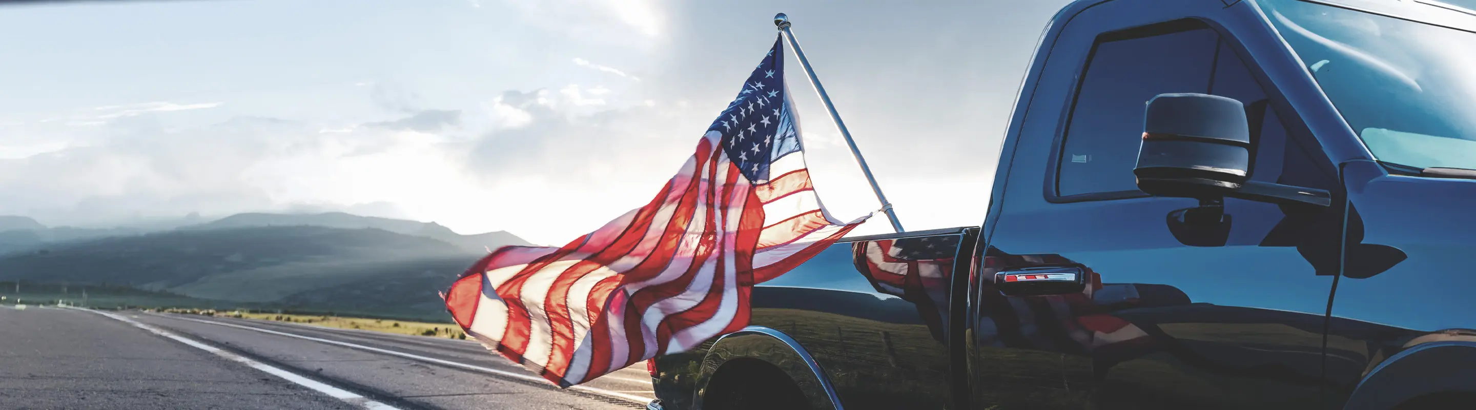A truck on an open road with an American flag waving from the back, set against a scenic mountain landscape under a cloudy sky.