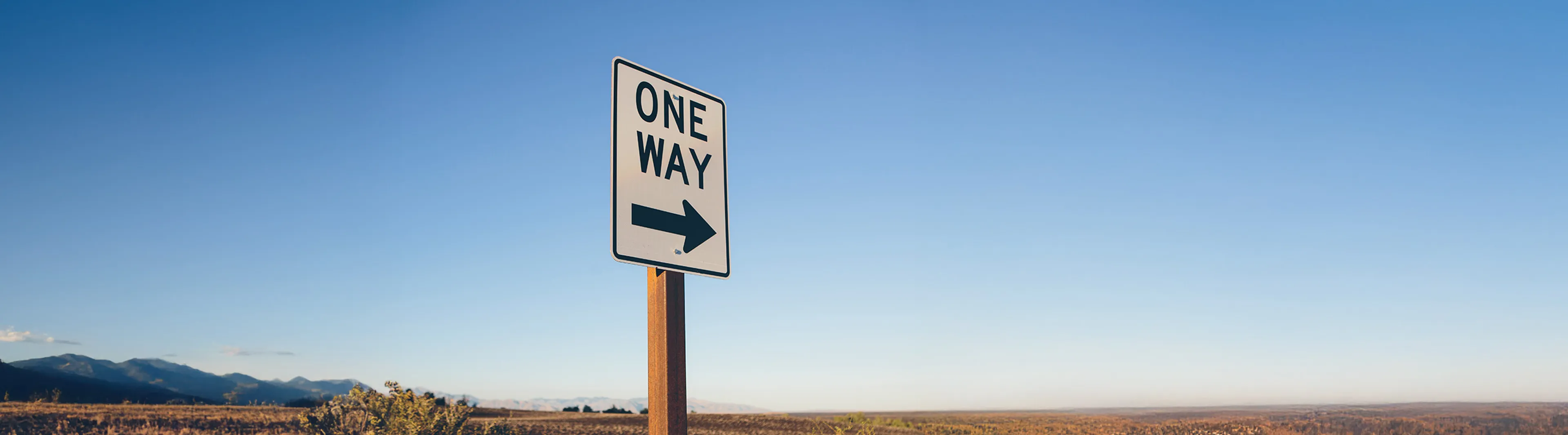 A "One Way" sign with an arrow pointing right stands against a clear blue sky, with distant mountains and open landscape in the background.