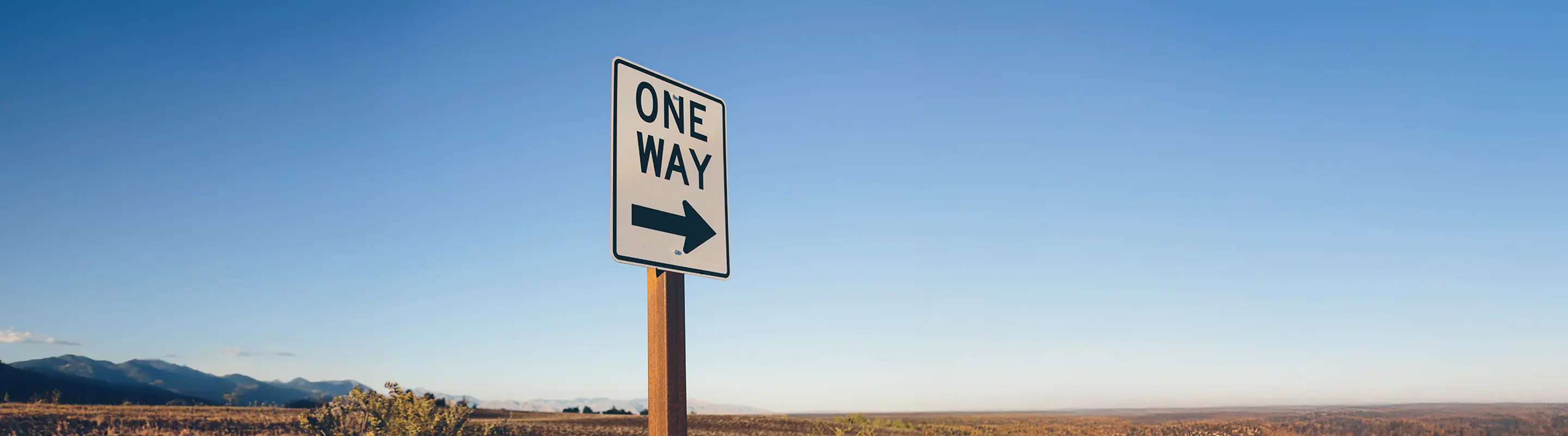 A "One Way" sign with an arrow pointing right stands against a clear blue sky, with distant mountains and open landscape in the background.