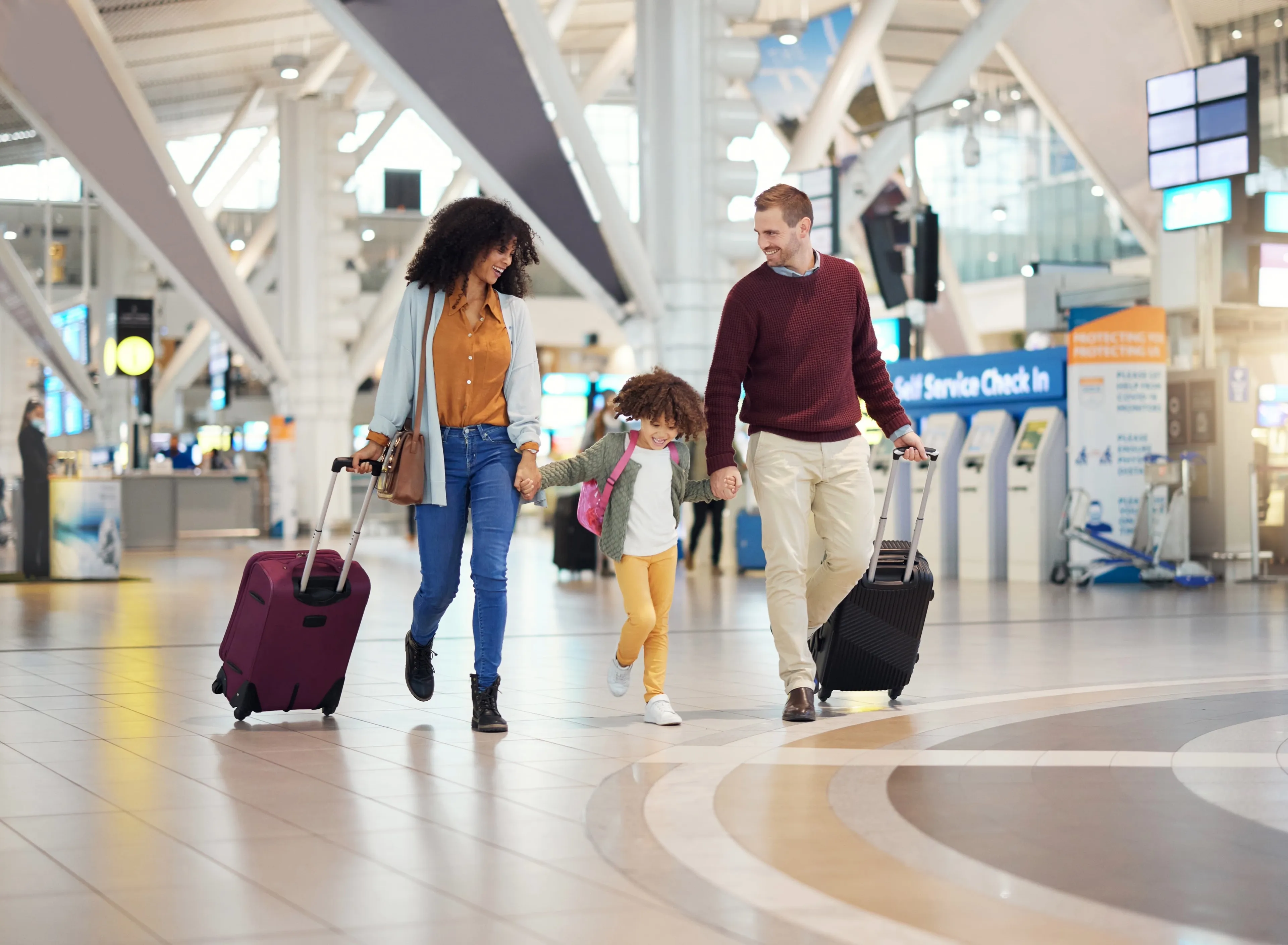 A family of three walks through an airport terminal, carrying luggage and smiling, with bright overhead lights and check-in counters in the background.