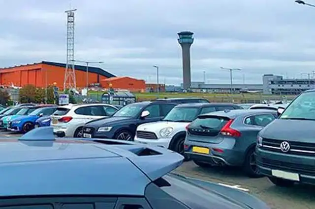 Cars parked near airport buildings with a control tower in the background.