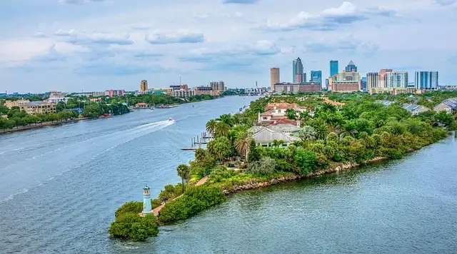 Aerial view of a river with boats near Tampa Bay and a city skyline in the background under a cloudy sky.