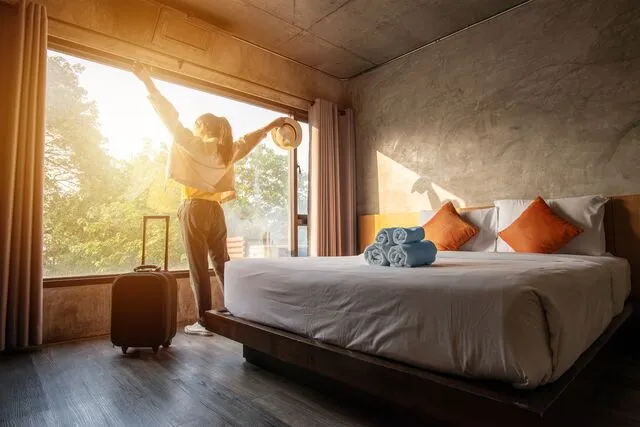 A person stands by a large window in a sunlit hotel room, stretching with arms raised, a suitcase nearby, and towels on the bed.