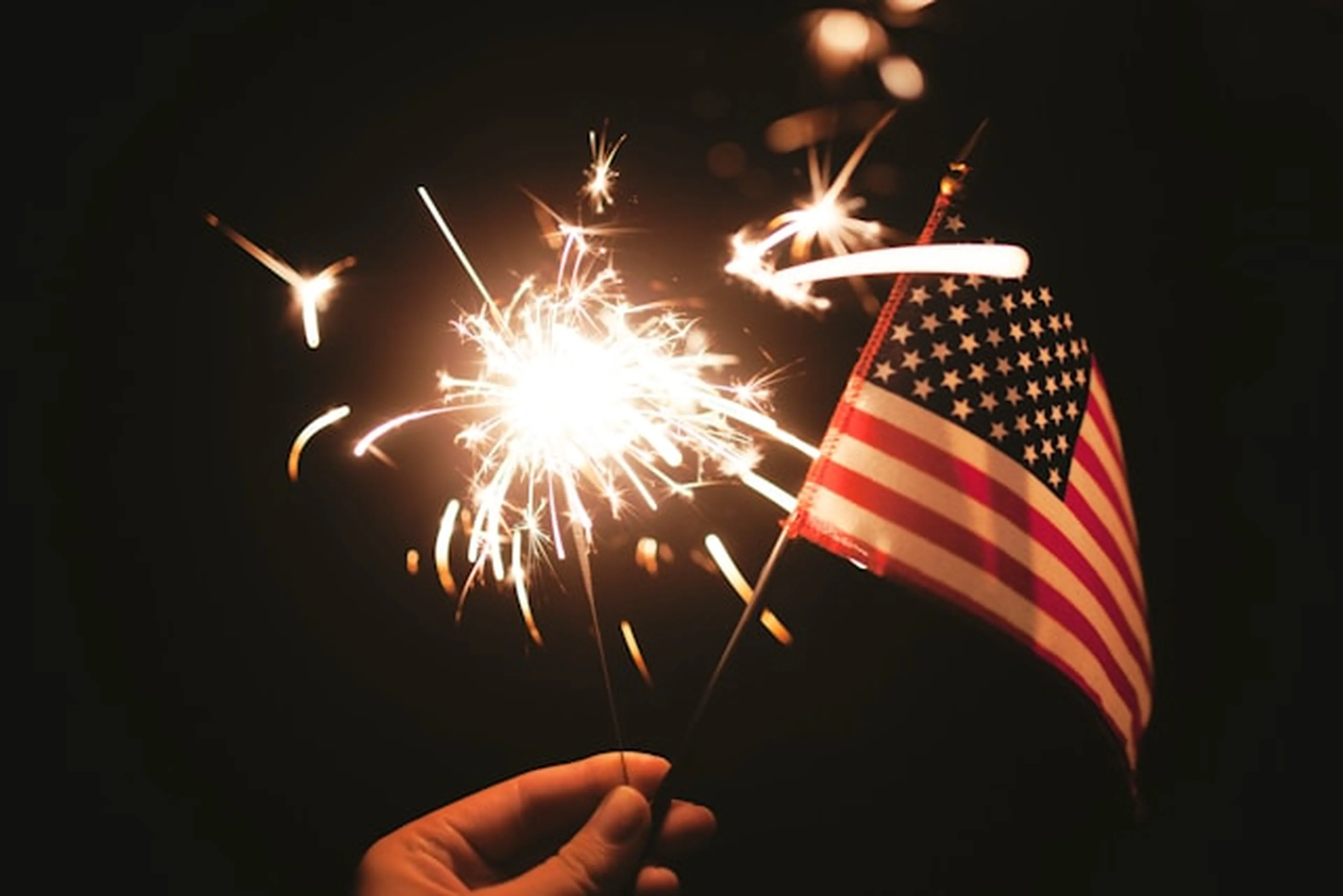 A hand holds a lit sparkler and a small American flag against a dark background.