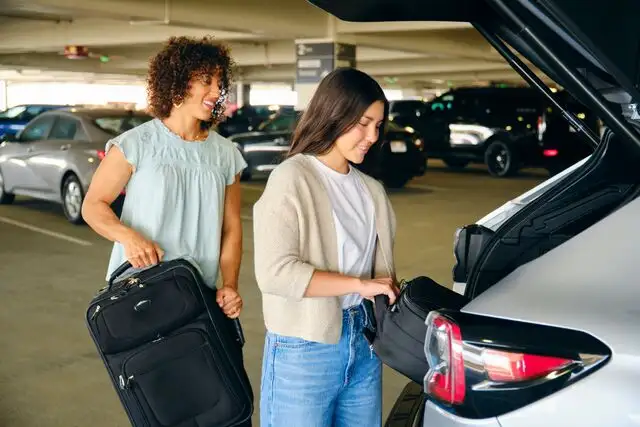 Two women loading luggage into the trunk of a silver car in a parking garage.
