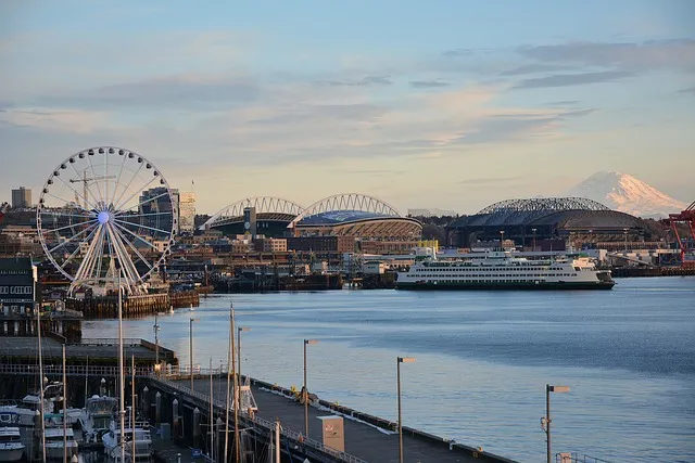 Seattle waterfront with a large ferris wheel, ferry, stadiums, and Mount Rainier in the distance under a blue sky.
