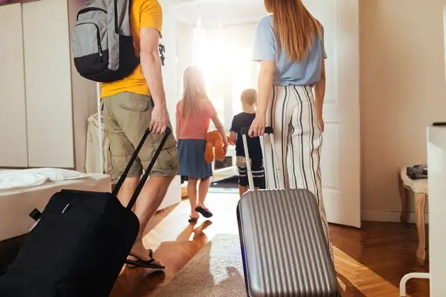 Family with luggage entering hotel room, two children carrying toys, and parents pulling suitcases. Bright, welcoming interior with wooden floor.