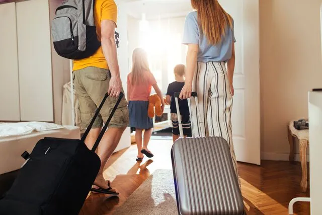 Family with luggage entering hotel room, two children carrying toys, and parents pulling suitcases. Bright, welcoming interior with wooden floor.