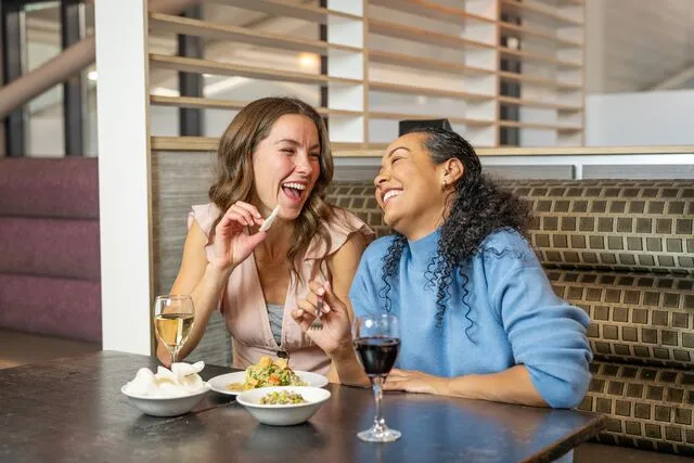 Two women laughing and enjoying drinks and snacks at a restaurant table, with wooden divider panels in the background.